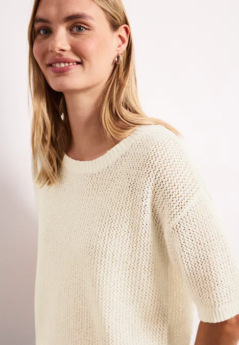 Junge Frau mit hellblondem Haar in einem cremefarbenen, kurzärmligen Strickpullover lächelt.