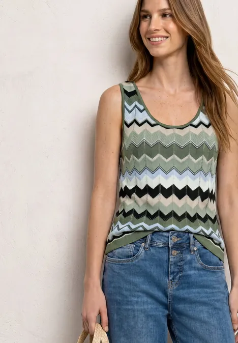 Vrouw in mouwloos top met groene-wit-zwarte zigzagprint en blauwe jeans, ze houdt een telefoon vast.