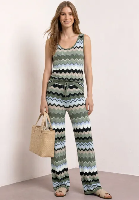 Vrouw in mouwloze jumpsuit met pastelgroen zigzagpatroon en rotan handtas.