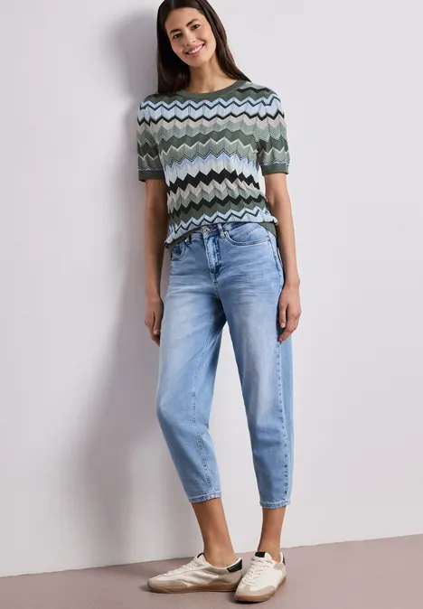 Frau in Zickzack-Pullover, hellblauer Jeans und Slipper vor neutralem Hintergrund.