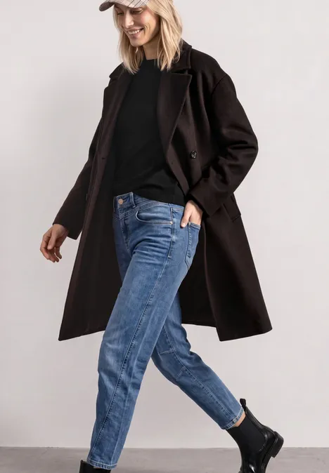 Frau in lässigem Outfit mit langem schwarzen Mantel, schwarzem Pullover, blauen Jeans, schwarzen Boots und weißer Mütze.