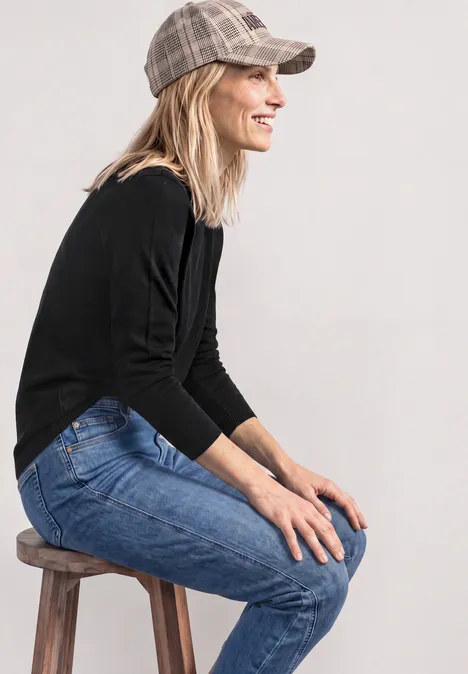 Frau sitzt lächelnd seitlich auf einem Hocker, trägt Jeans, schwarzes Shirt und Schirmmütze.