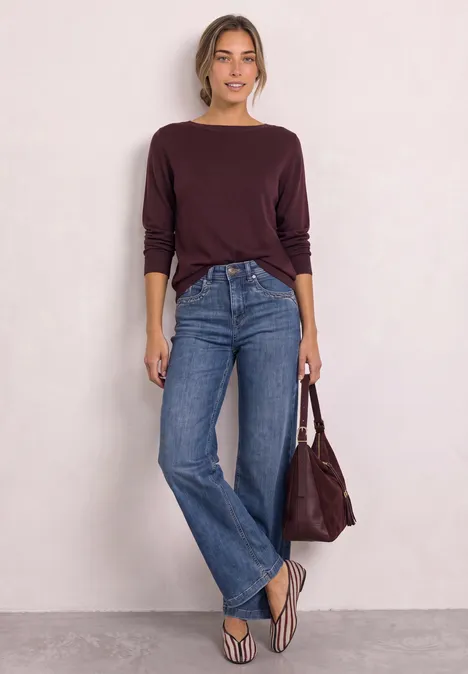 Frau in weinrotem Pullover, blauen Jeans, Flip-Flops, weiße Wand, hält passende Handtasche.