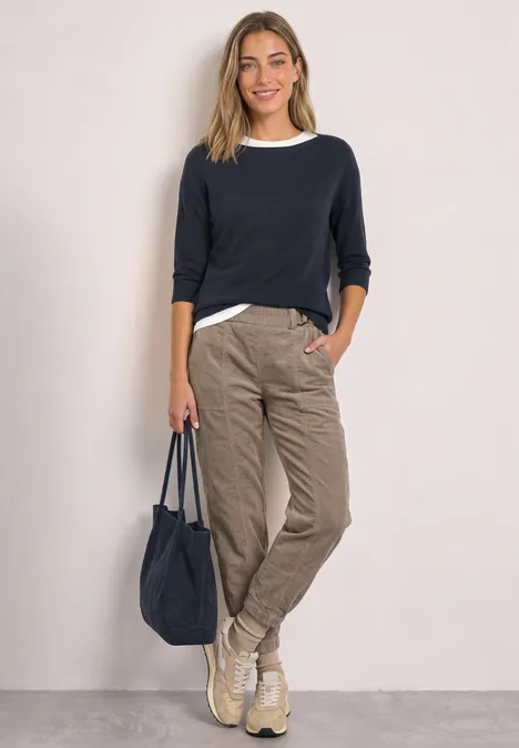 Frau in dunkelblauem Pullover, weißem Shirt, beige Cargohose, Sneakern und passender Tasche