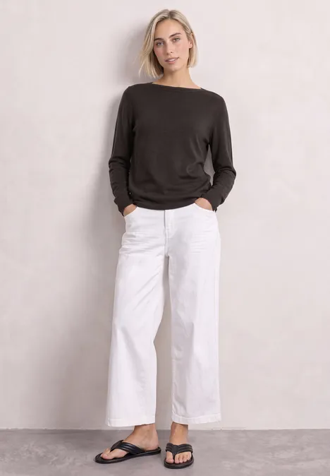 Eenvoudige trui met boothals black coffee Frau in schwarzem Pullover, weißer weiter Hose und schwarzen Sandalen vor neutralem Hintergrund.