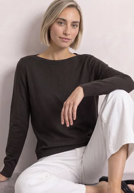 Eenvoudige trui met boothals black coffee Frau mit kurzem blondem Bob in schwarzem Pullover und weißen Hosen sitzt entspannt vor neutralem Hintergrund.