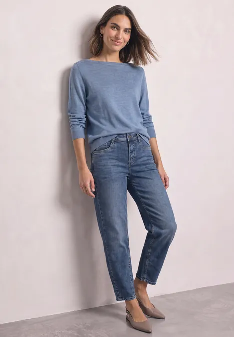Pull basique à col bateau northern blue mel. Femme en pull bleu et jean, debout devant un mur clair.