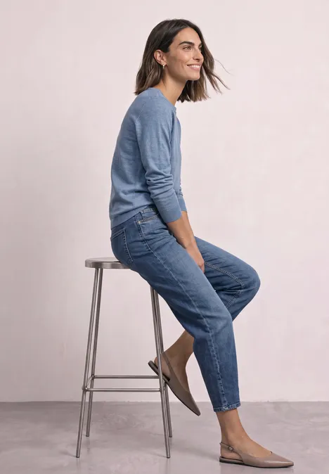 Pull basique à col bateau northern blue mel. Femme assise de profil sur un tabouret, pull bleu, jean et chaussures plates, fond clair.