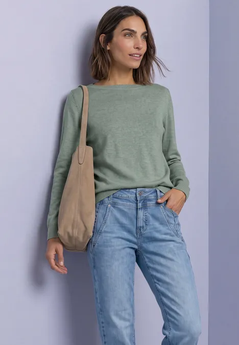 Eenvoudige trui met boothals eucalyptus green mel. Eine Frau trägt grünen Pullover, blaue Jeans und eine beige Schultertasche.