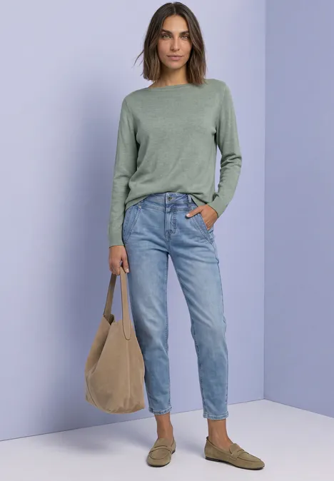 Eenvoudige trui met boothals eucalyptus green mel. Frau in grünem Pullover, Jeans und braunen Slippern trägt eine beige Tasche vor lilafarbenem Hintergrund.