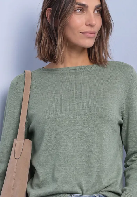 Eenvoudige trui met boothals eucalyptus green mel. Frau mit schulterlangem braunem Haar, trägt grünen Pullover und beige Umhängetasche vor hellem Hintergrund.