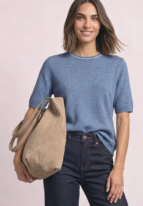 Pull à manches courtes avec détails contrastés northern blue mel. Eine Frau im blauen kurzärmligen Pullover und dunklen Jeans trägt lächelnd eine große beige Tasche.