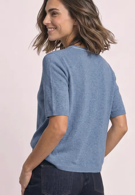 Pull à manches courtes avec détails contrastés northern blue mel. Frau mit braunen, gewellten Haaren im blauen Kurzarmshirt und Jeans, die lächelnd über die Schulter blickt.