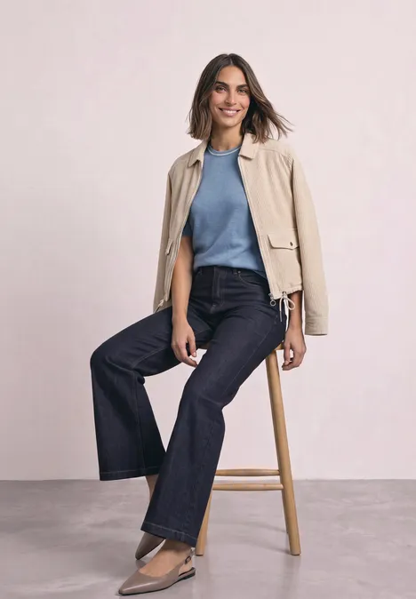 Pull à manches courtes avec détails contrastés northern blue mel. Eine Frau in beiger Jacke, blauem Pullover und weitem Jeans sitzt lächelnd auf einem Hocker.