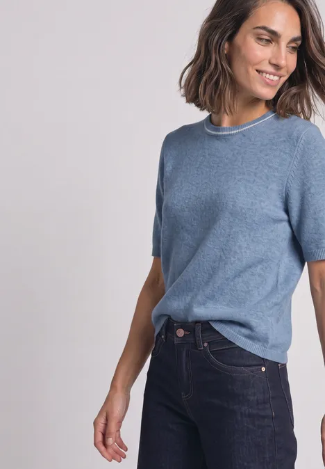 Pull à manches courtes avec détails contrastés northern blue mel. Frau in hellblauem Kurzarm-Pullover und dunkler Jeans lächelt seitlich.