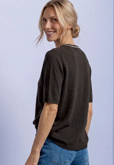 Pull à manches courtes avec détails contrastés black coffee mel. Lächelnde Frau mit locker zusammengebundenen blonden Haaren blickt über die Schulter, trägt dunkles Shirt und Jeans.