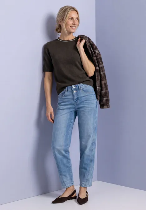 Pull à manches courtes avec détails contrastés black coffee mel. Frau lehnt an lila Wand, trägt schwarzes Shirt, blaue Jeans und schwarze Ballerinas, eine karierte Jacke über der Schulter.