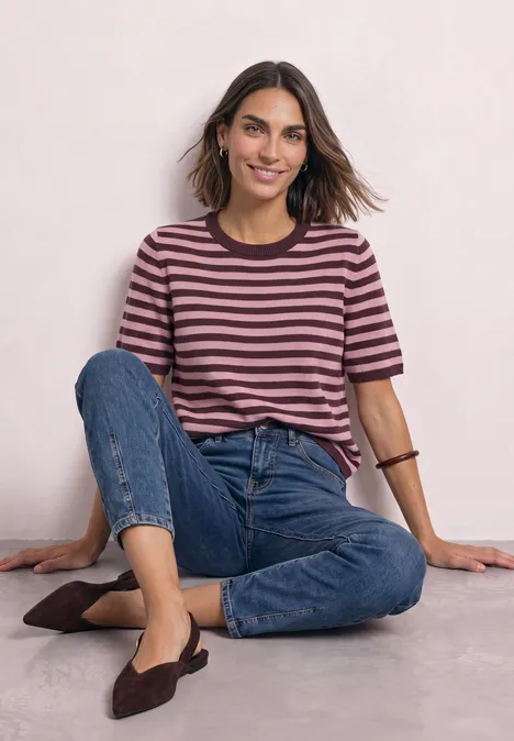T-shirt met korte mouwen, ronde hals en strepen rustic rouge mel. Frau sitzt lächelnd im Schneidersitz, trägt gestreiftes Shirt, Jeans und flache Schuhe vor hellem Hintergrund.