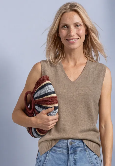 V-hals trui in effen kleur sanded amber mel. Frau mit blondem Haar, trägt einen ärmellosen Pullover, Jeans und hält eine farbige gestreifte Tasche.