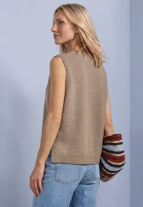 V-hals trui in effen kleur sanded amber mel. Eine blonde Frau trägt eine beige, ärmellose Strickweste, blaue Jeans und hält eine bunte, gestreifte Tasche.
