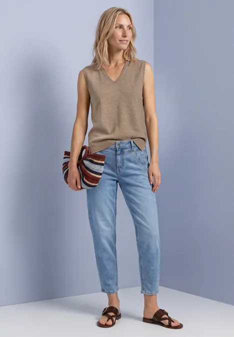 V-hals trui in effen kleur sanded amber mel. Frau in ärmellosem, beigefarbenem V-Pullover, heller Jeans und schwarzen Sandalen, mit gestreifter Tasche.