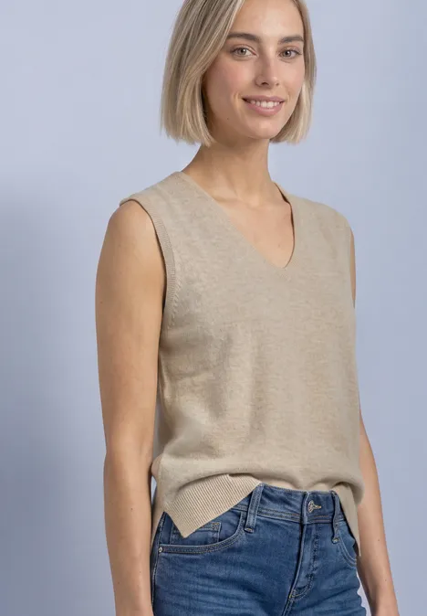 V-hals trui in effen kleur light sepia sand mel. Junge Frau mit hellblondem Bob, trägt einen beigen V-Ausschnitt-Pulli ohne Ärmel und blaue Jeans, lächelnd.