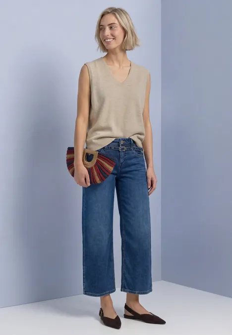 V-hals trui in effen kleur light sepia sand mel. Frau vor hellviolettem Hintergrund trägt beiges ärmelloses Top, weite Cropped-Jeans, flache Schuhe und runde Tasche.