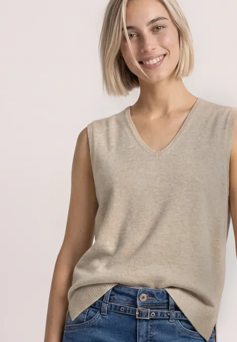 V-hals trui in effen kleur light sepia sand mel. Frau trägt ärmellosen, beigefarbenen V-Ausschnitt-Strickpullunder zu blauen Jeans.