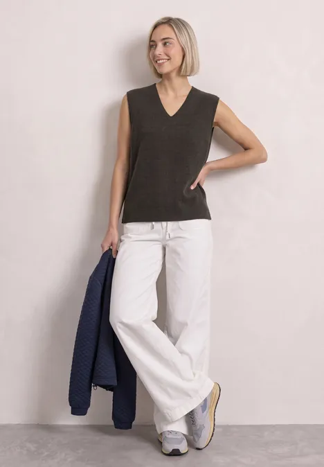 V-hals trui in effen kleur black coffee mel. Frau mit blondem Bob trägt ärmelloses schwarzes Top, weiße Schlaghose und Sneaker, hält blauen Blazer.
