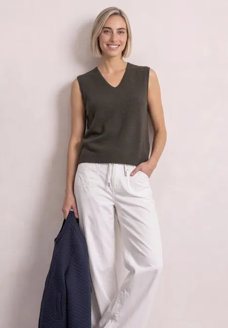 V-hals trui in effen kleur black coffee mel. Eine lächelnde Frau in dunkelgrauem Top und weißen Hosen hält eine blaue Jacke.
