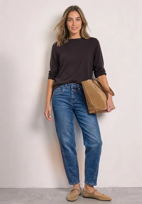 Basic Pullover mit U-Boot-Ausschnitt ruby vine Frau in schwarzem Langarmshirt, blauen Jeans und beigen Slippern steht lächelnd vor neutralem Hintergrund und hält Tasche.