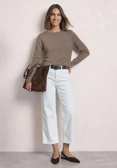 Eine lässige Frau in braunem Pullover, weißen Capri-Hosen und brauner Schultertasche vor neutralem Hintergrund.