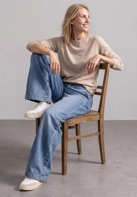 Eenvoudige trui met boothals clay sand mel. Eine lächelnde Frau sitzt entspannt auf einem Holzstuhl, trägt beigen Pullover, weite Jeans und weiße Sneaker.