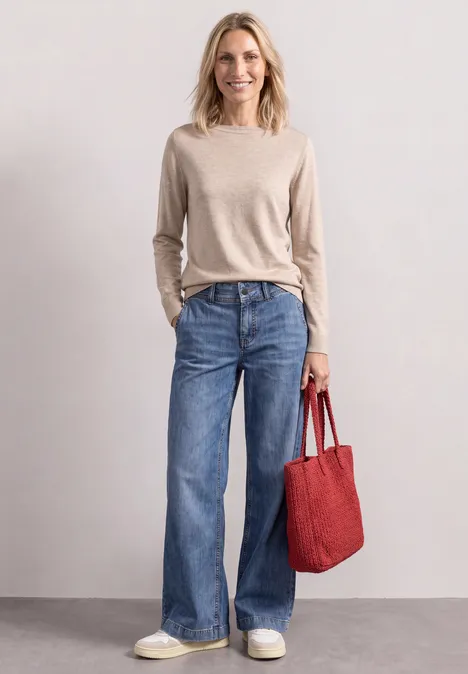 Eenvoudige trui met boothals clay sand mel. Eine Frau in hellem Pullover und weiten Jeans trägt eine rote Tasche und weiße Sneaker.