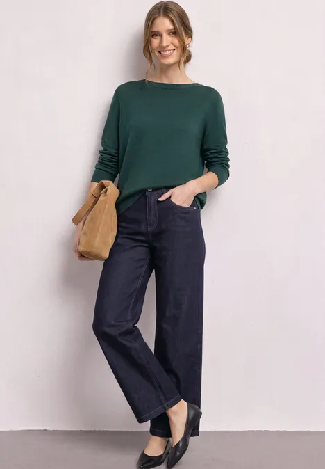 Eenvoudige trui met boothals shadow teal Een lachende vrouw draagt een groene trui, wijde spijkerbroek, zwarte schoenen en houdt een bruine clutch vast.