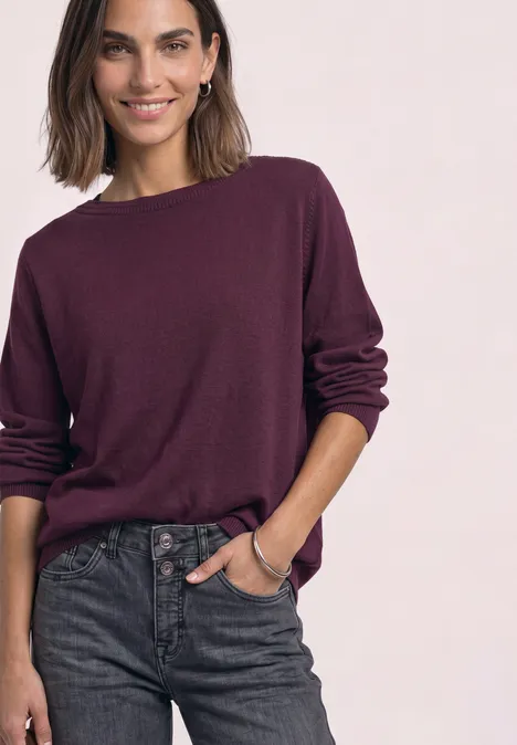 Eenvoudige trui met boothals berry maroon Vrouw met paarse trui en donkere jeans voor lichte achtergrond.