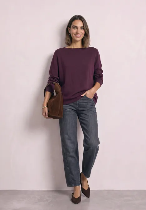 Eenvoudige trui met boothals berry maroon Vrouw in paarse trui, grijze jeans en donkere instappers, met bruine tas voor een lichte muur.
