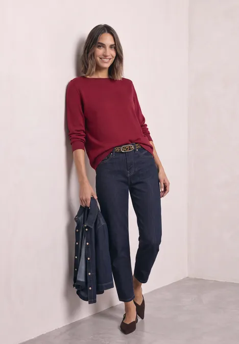 Eenvoudige trui met boothals inferno red Vrouw leunt lachend tegen lichte muur, draagt bordeaux trui, donkere jeans, riem en spijkerjack