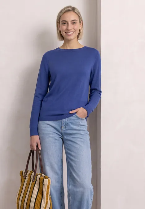 Eenvoudige trui met boothals bold blue Een vrouw voor een lichte achtergrond draagt een blauw shirt, spijkerbroek en een gestreepte tas.