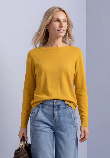Eenvoudige trui met boothals saffron yellow Eine Frau mit senfgelbem Pullover und blauen Jeans hält eine Handtasche und blickt seitlich.