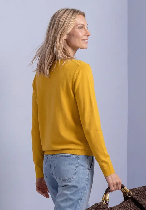 Eenvoudige trui met boothals saffron yellow Eine Frau steht seitlich, trägt einen gelben Pullover, blaue Jeans und hält eine braune Tasche vor hellem Hintergrund.