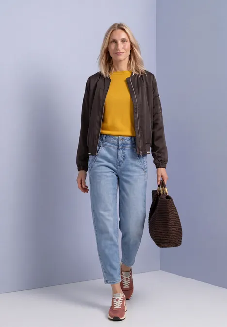 Eenvoudige trui met boothals saffron yellow Frau in gelbem Shirt, dunkler Jacke, Jeans und Sneakers trägt eine braune Tasche vor hellblauem Hintergrund