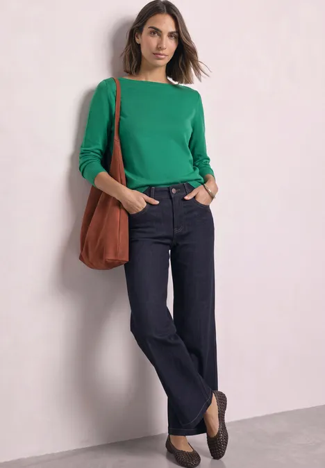 Eenvoudige trui met boothals kelly green Vrouw draagt groen lang shirt, donkere jeans, bruine schoudertas en loafers, leunt tegen een muur.