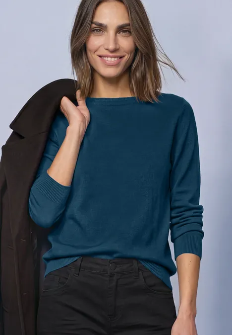 Frau in blauem Pullover und schwarzer Hose trägt Jacke über der Schulter und lächelt.