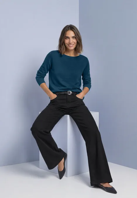 Frau in blaugrünem Pullover und schwarzen, weiten Hosen, lächelnd in minimalistischem Raum posierend.