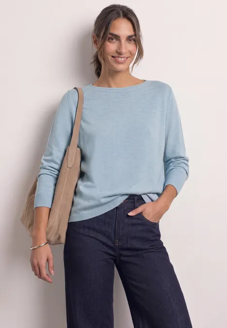 Eenvoudige trui met boothals pale blue mel. Een glimlachende vrouw in lichtblauwe trui en donkere jeans, met een schoudertas voor een lichte achtergrond.
