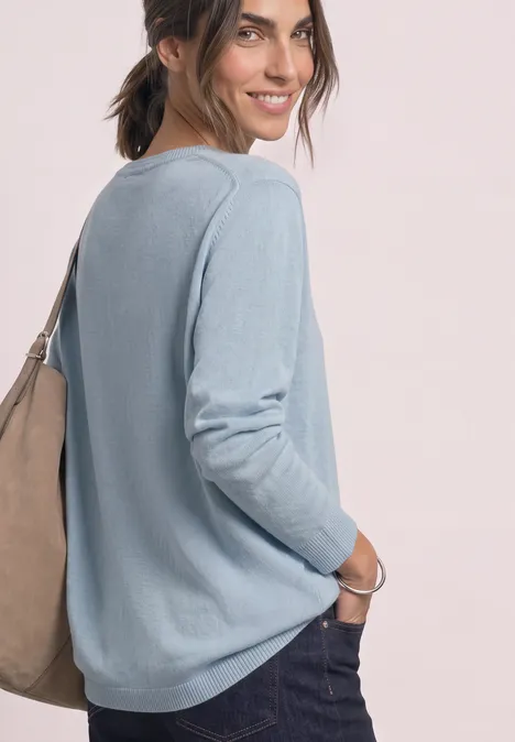 Eenvoudige trui met boothals pale blue mel. Vrouw glimlachend in een lichtblauwe trui en jeans, zijaanzicht met een schoudertas, relaxte houding.