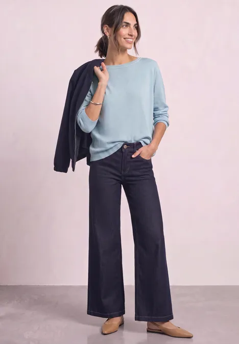 Eenvoudige trui met boothals pale blue mel. Vrouw in lichtblauwe trui en wijde jeans, nonchalant met een donker jasje over de schouder.