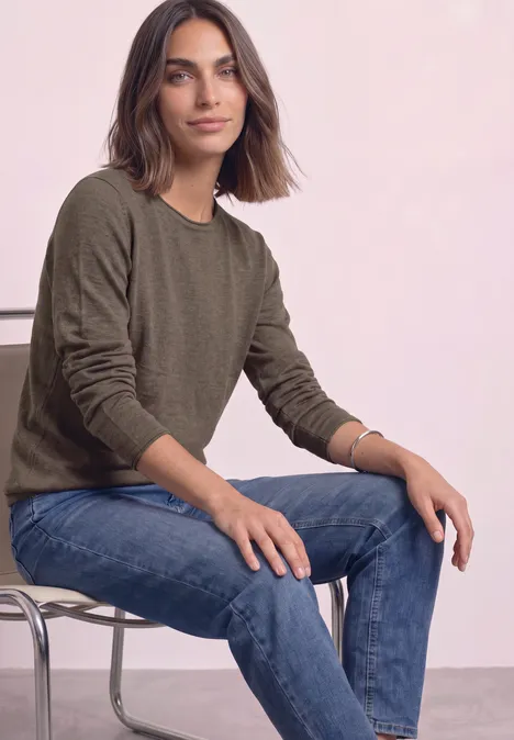 Trui met ronde hals en omgeslagen randen nomad brown mel. Een vrouw met bruin haar zit ontspannen op een stoel, draagt een groene trui en een blauwe jeans.
