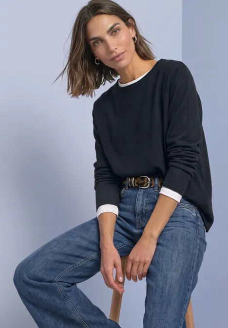 Frau in schwarzem Pullover mit weißem Kragen und blauen Jeans sitzt lässig auf einem Hocker vor grauem Hintergrund.